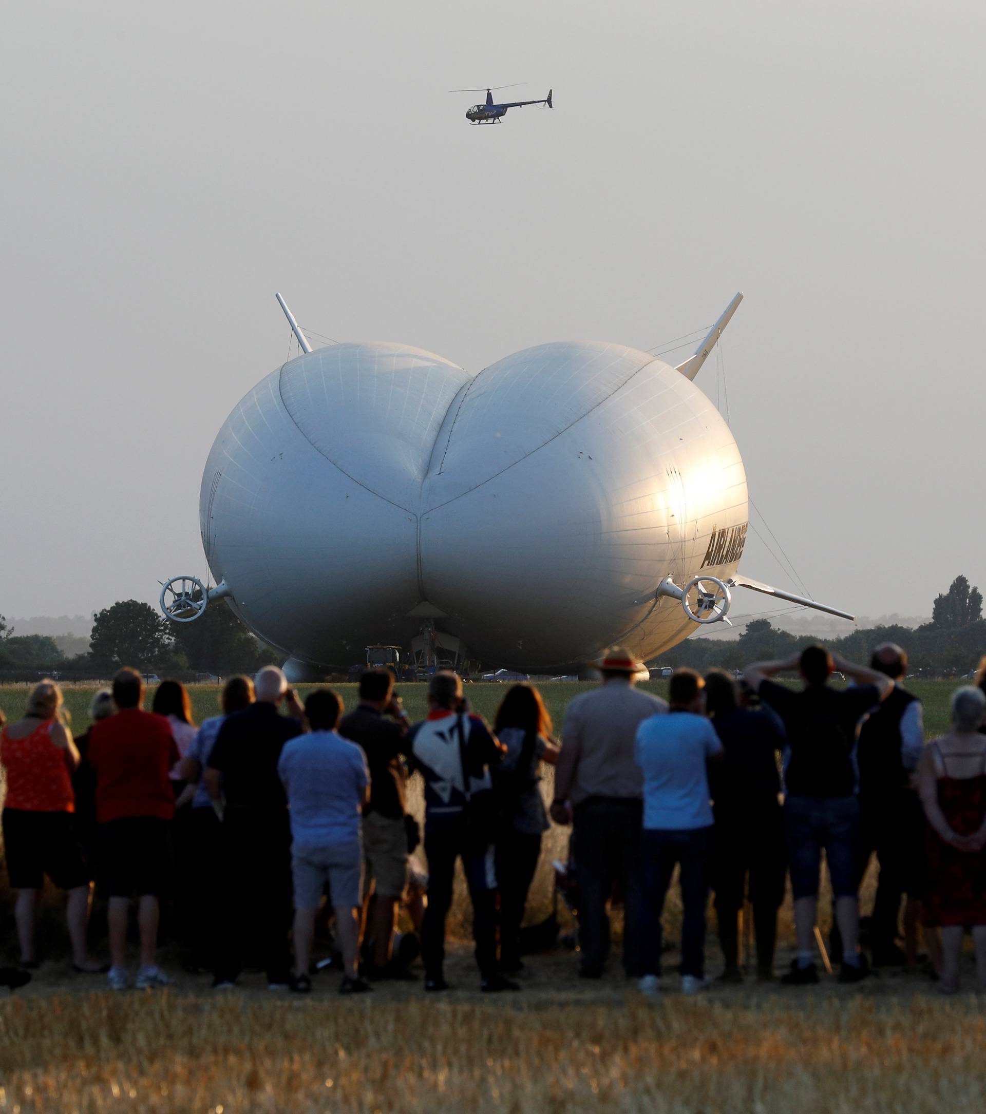 A helicopter flies over the Airlander 10 hybrid airship before its maiden flight at Cardington Airfield in Britain