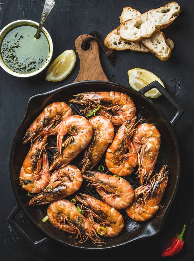 Roasted tiger prawns in iron grilling pan on wooden board with fresh leek, lemon slices, pepper, bread and pesto sauce over black background