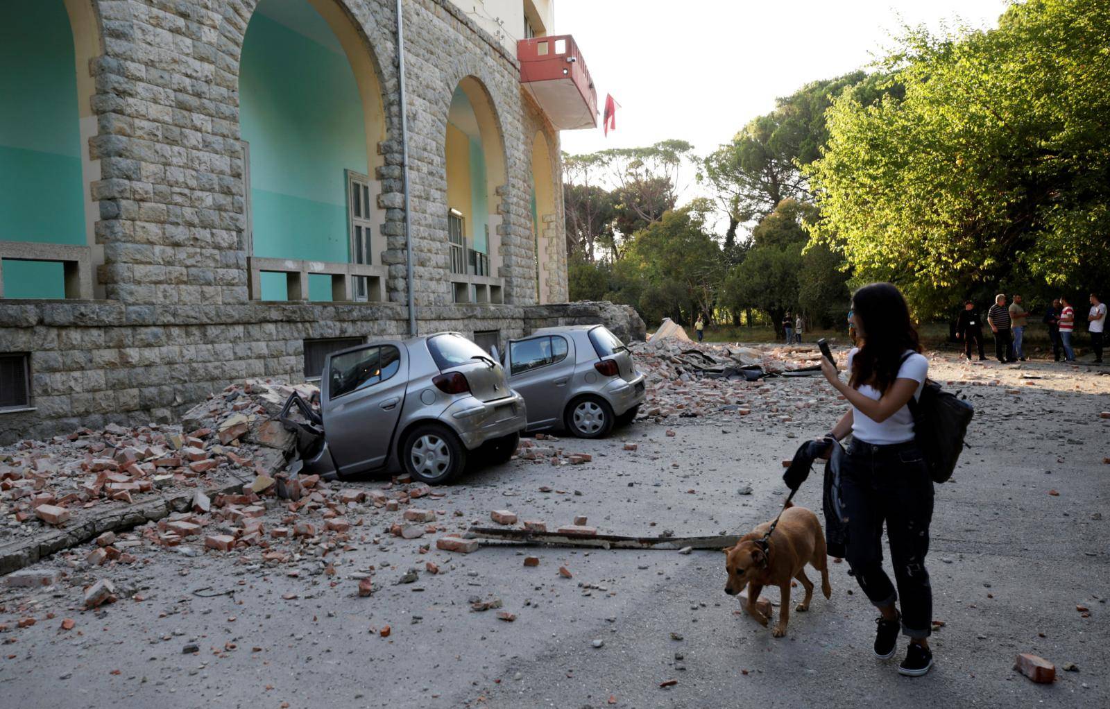 A woman with a dog walks past destroyed cars and a damaged building after an earthquake in Tirana