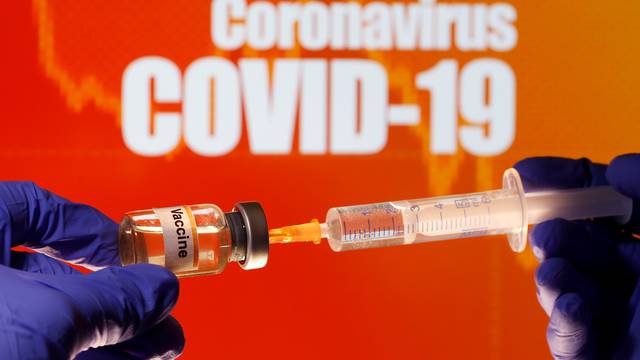 A small bottle labeled with a "Vaccine" sticker is held near a medical syringe in front of displayed "Coronavirus COVID-19" words in this illustration