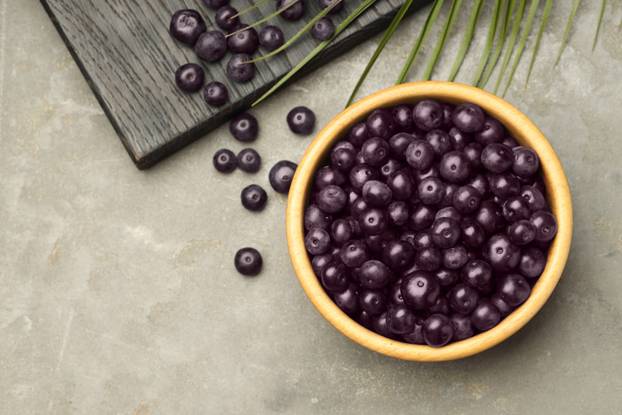 Ripe acai berries in bowl on grey textured table, flat lay. Spac