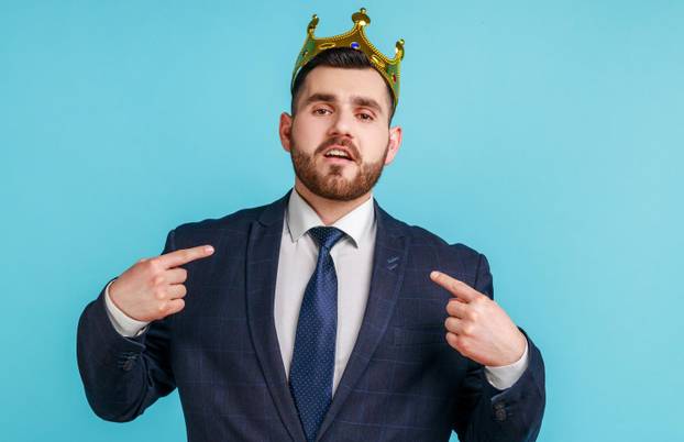 Proud bearded man wearing official style suit egoistically looking at camera, posing with crown on head, pretending to be king, pointing at himself.