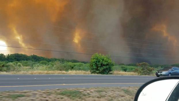 A thick plume of smoke hovers over a wildfire next to a road in Maui