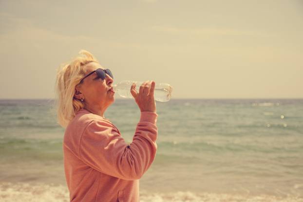 Adult blonde woman drinks water from a bottle with the sea in the background