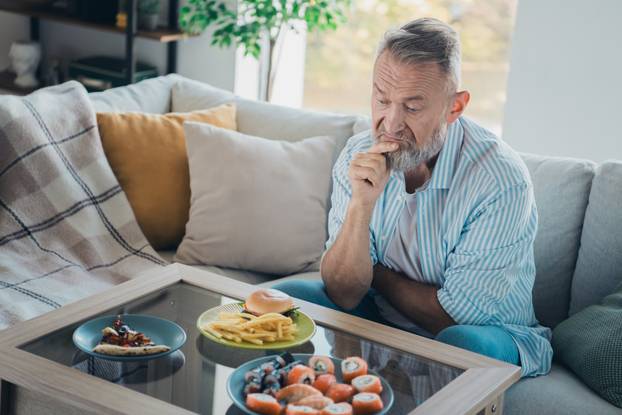 Elderly man in casual striped shirt thoughtfully contemplating food options while seated in a cozy, well-decorated living room