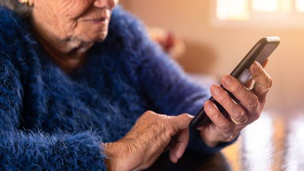 Elderly woman using mobile phone while sitting at living room home. Grandmother