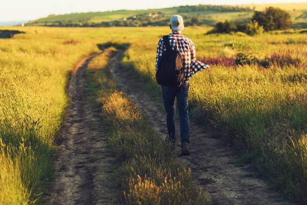 Active tourist, a man with a briefcase in nature, a hipster in a cap is walking along a path on a green hill, healthy lifestyle