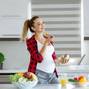 Pregnant blond young woman in red checkered shirt at kitchen eating a fruit salad, smiling, looking away, in front of her bowls with fresh vegetables, juice and fruits. Healthy eeating concept