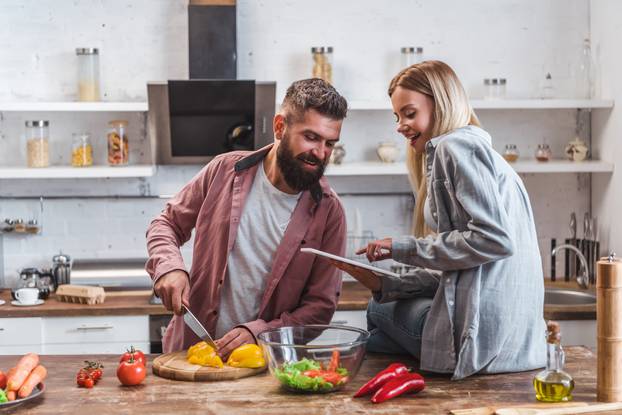 Husband making dinner and wife sitting on table with digital tablet 