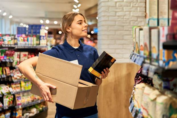 Young female store worker stocking shelves in a supermarket aisle. She holds a cardboard box and a packaged item, focusing on organizing the products.