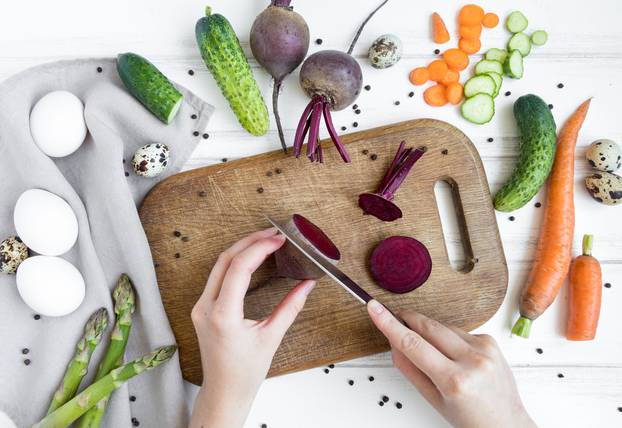 Woman hands slicing beetroot on wooden cutting board, surrounded by vegetables. Flat lay, top view