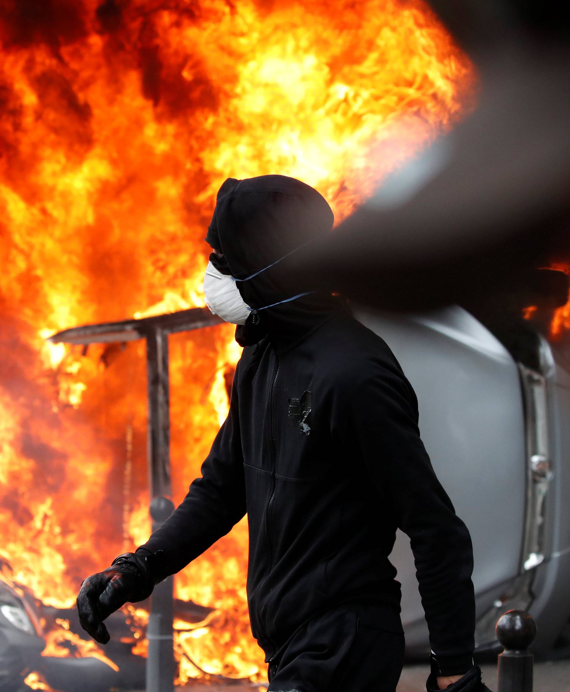 A masked protester walks near a car that burns outside a Renault automobile garage during clashes during the May Day labour union march in Paris