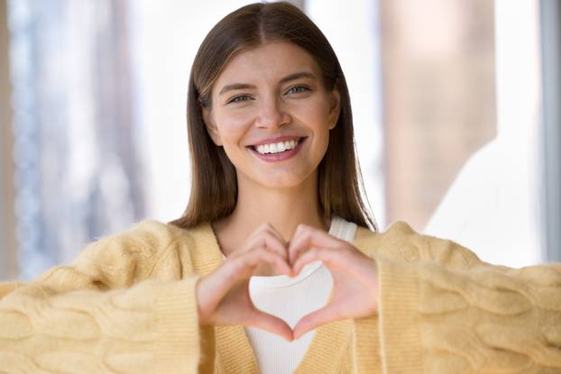 Cheerful beautiful young woman keeping finger heart at chest