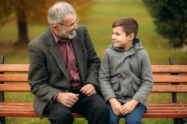 Grandpa and his grandson spend time together in the park. They are sitting on the bench and look to each other