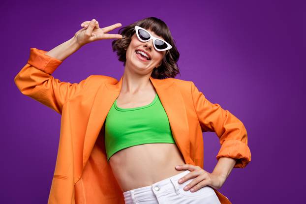 Photo of sweet adorable young lady wear orange jacket dark glasses smiling showing v-signs isolated violet color background