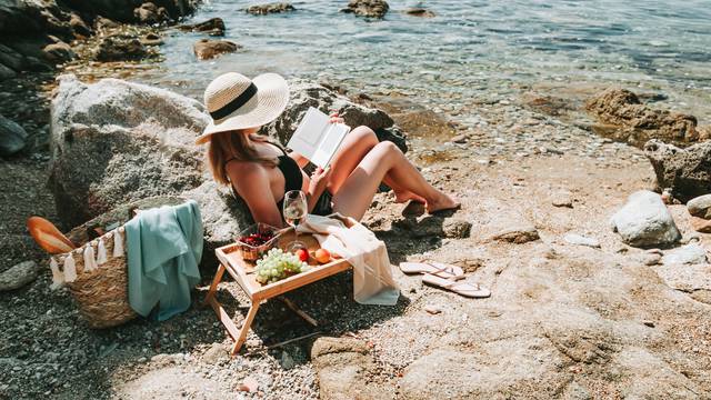 Woman in bikini and hat alone at beach reading electronic book,