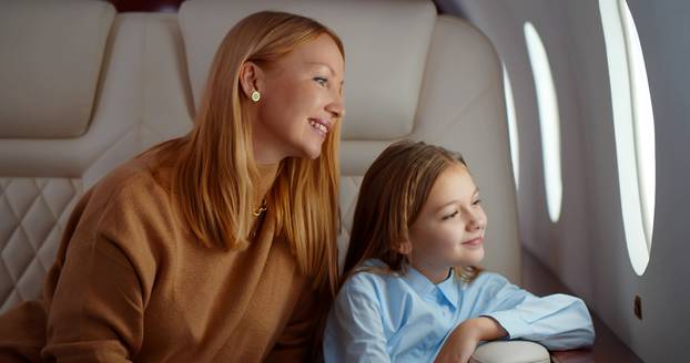 Mature elegant mother and preteen daughter looking through window flying on airplane