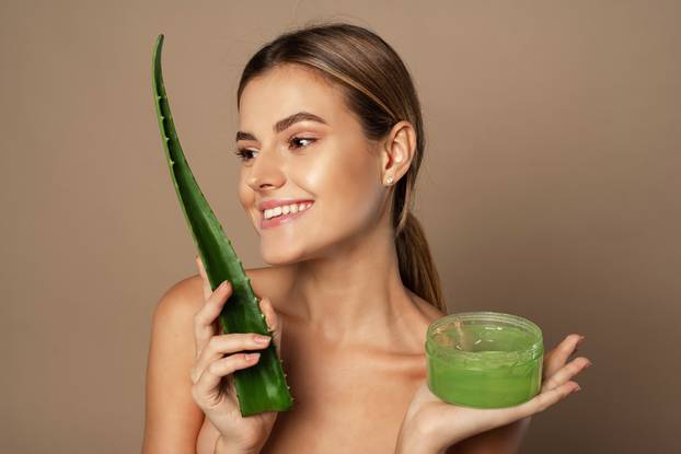 Smiling happy young female model holding aloe leaf and jar of aloe gel on beige background. The concept of skin care, moisturizing with natural cosmetics