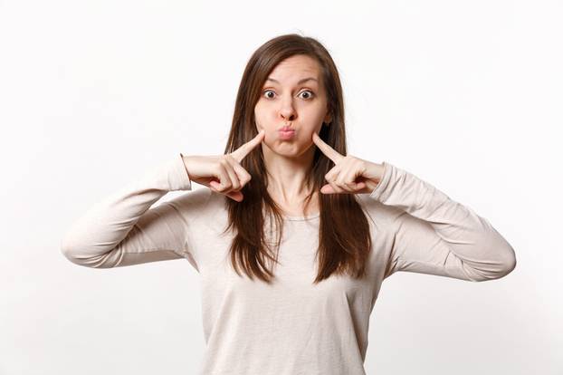 Portrait of funny young woman in light clothes pointing index fingers on blowing cheeks isolated on white wall background in studio. People sincere emotions, lifestyle concept. Mock up copy space.