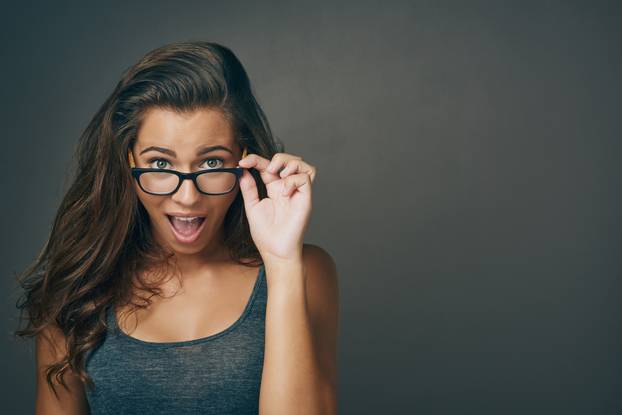 I cant believe it. Studio shot of an attractive young woman peering over her glasses against a grey background.