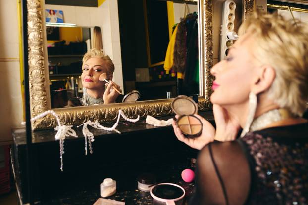 A drag queen artfully applies makeup in front of a mirror, readying for a festive show.