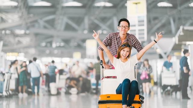 Young Asian tourist couple happy and excited together for the trip, girlfriend sitting and cheering on baggage trolley or luggage cart. Holiday vacation travelling abroad concept, with copy space