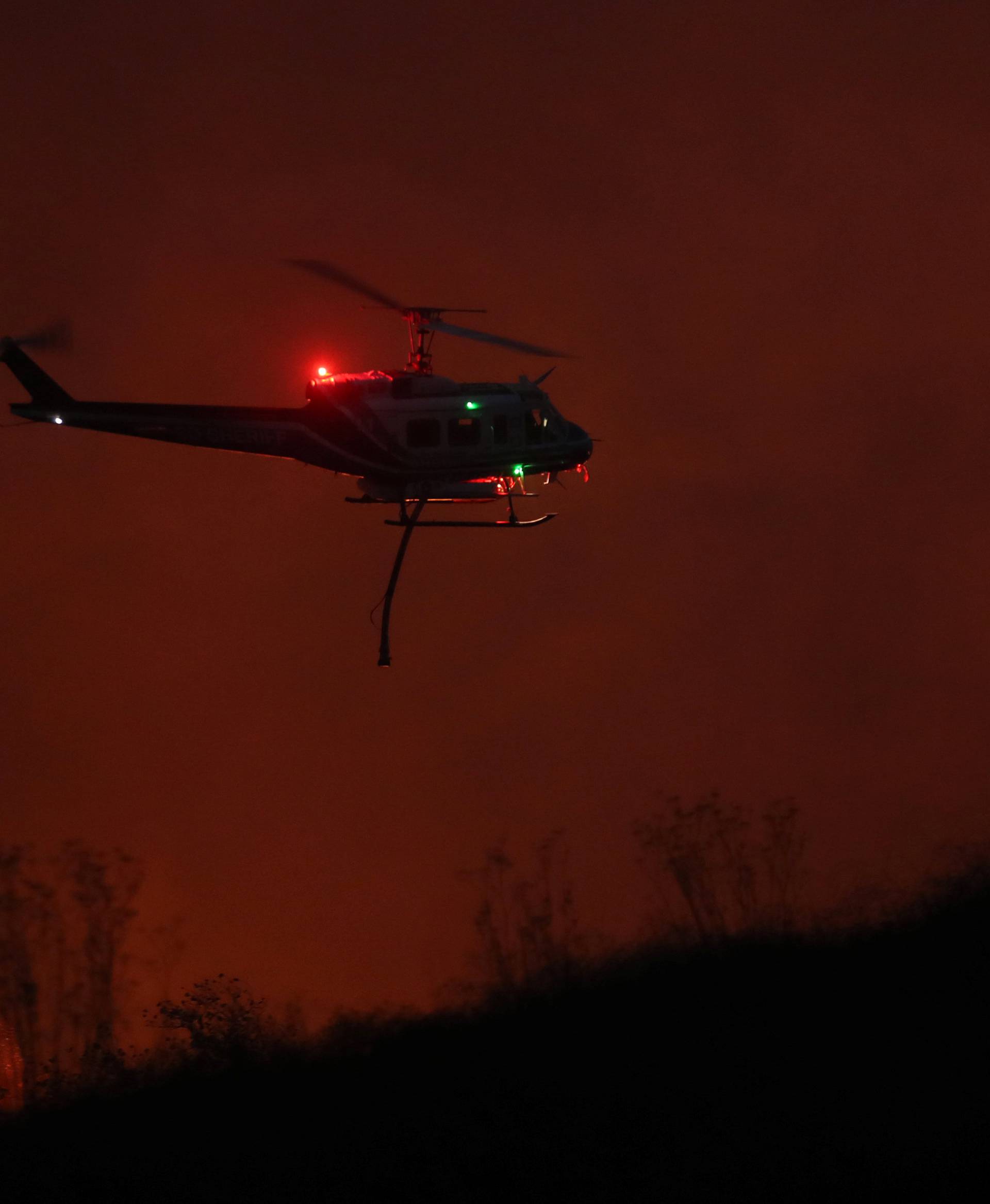 A fire fighting helicopter travels over the Lilac Fire as evening comes to Bonsall, California