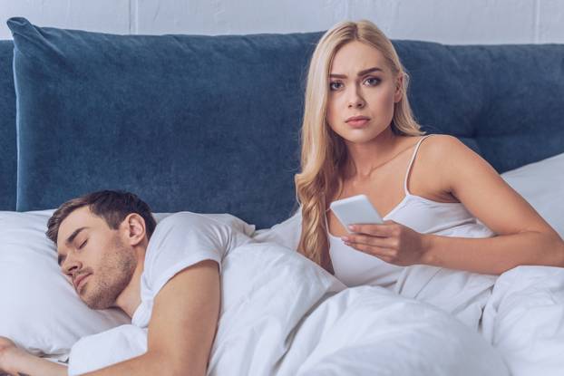 unhappy young woman holding smartphone and looking at camera while boyfriend sleeping in bed, distrust concept