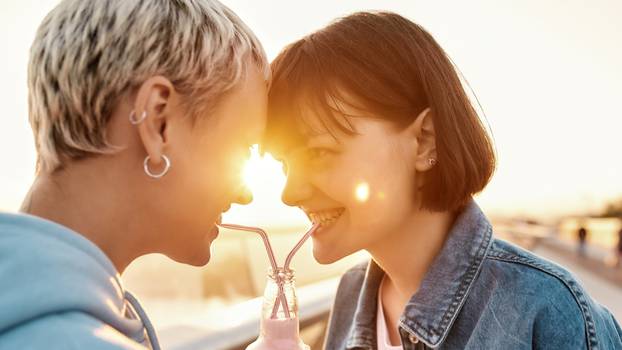 Close up of happy young lesbian couple drinking from one glass bottle with the straw, Two women enjoying cold beverage on a summer day outdoors