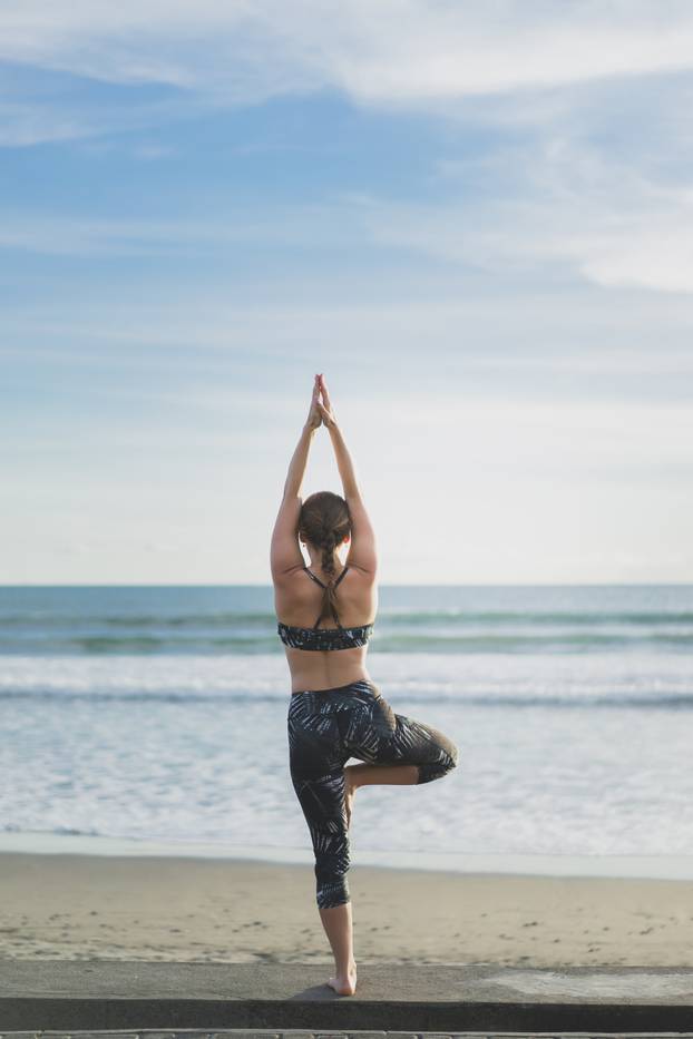 back view of woman practicing yoga in tree pose with ocean and blue sky on background, Bali, Indonesia