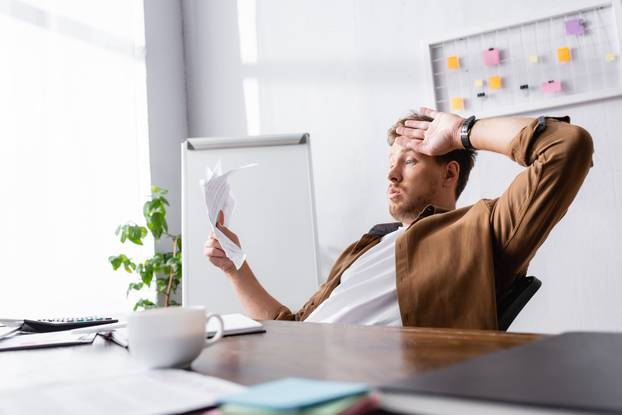 Selective focus of tired businessman holding papers near coffee cup on table in office 