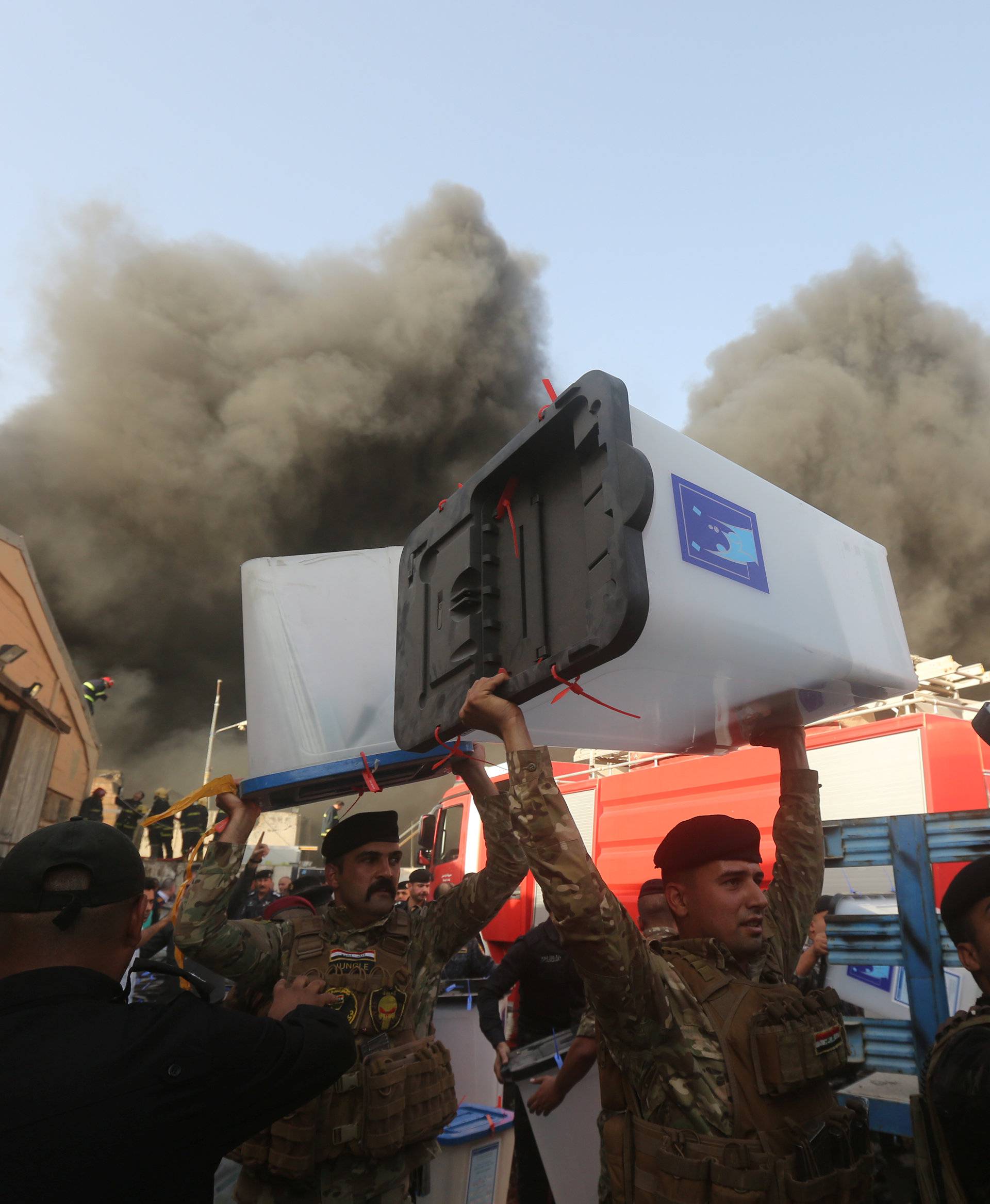Security forces carry ballot boxes as smoke rises from a storage site in Baghdad, housing the boxes from Iraq's May parliamentary election