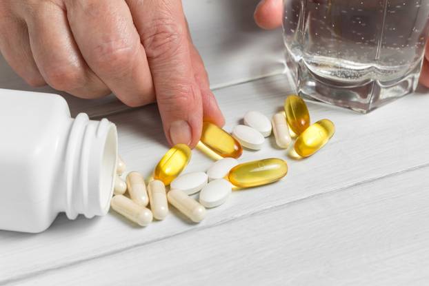 Woman hands takes yellow capsules of omega 3, white pills of calcium, glucosamine supplements, glass of water and plastic bottle on wooden planks table