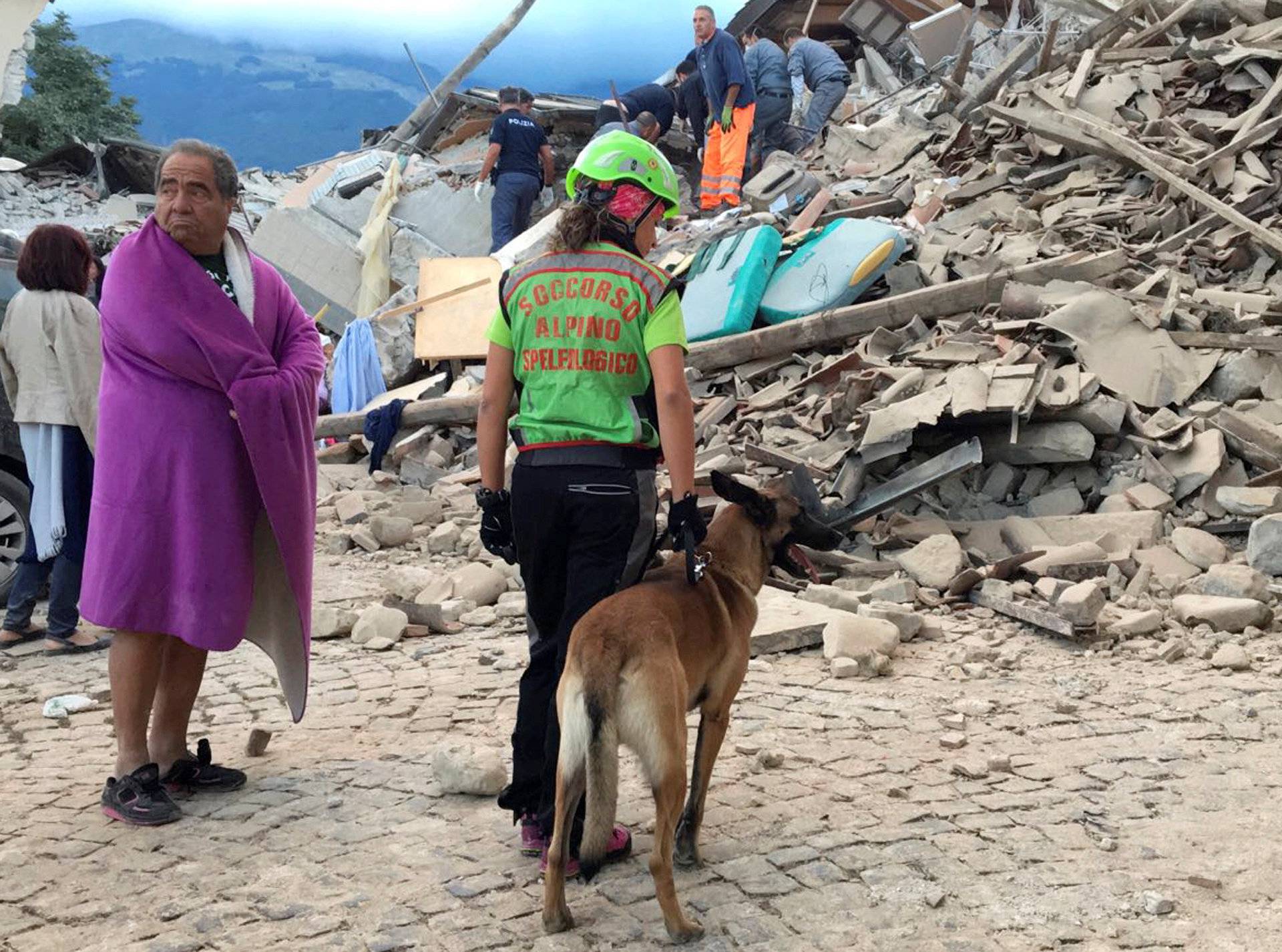 A man wrapped in blanket looks on as a rescuer with a dog stand in front a collapsed house following a quake in Amatrice 