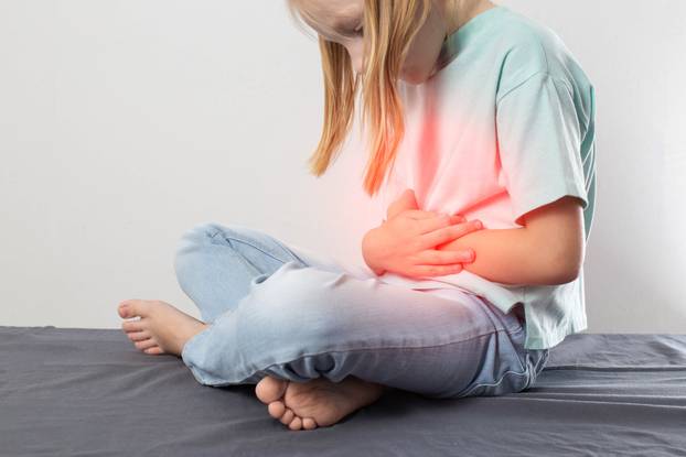 A seven-year-old blonde girl sits and holds her red belly. Concept of pain and cramps in the abdomen. Stomach diseases, gastritis and peptic ulcers in children. Copy space for text, dysfunction