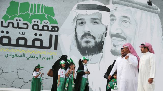 Saudi people walk past a poster depicting Saudi Arabia's King Salman bin Abdulaziz and Crown Prince of Saudi Arabia Mohammad bin Salman during the 89th annual National Day of Saudi Arabia in Riyadh