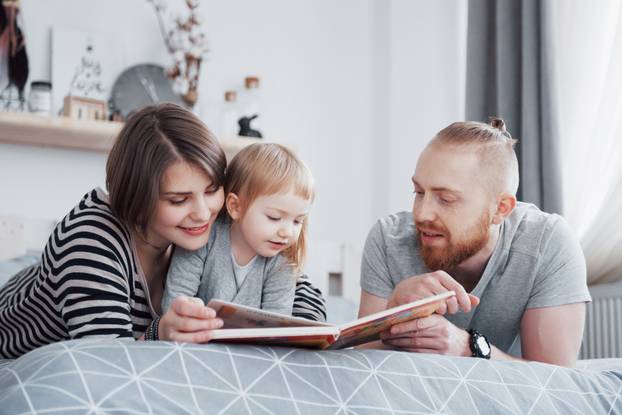 Father, Mother and Little Daughter Reading Children's Book on a Sofa in the Living Room. Happy big family read an interesting book on a festive day. Parents love their children