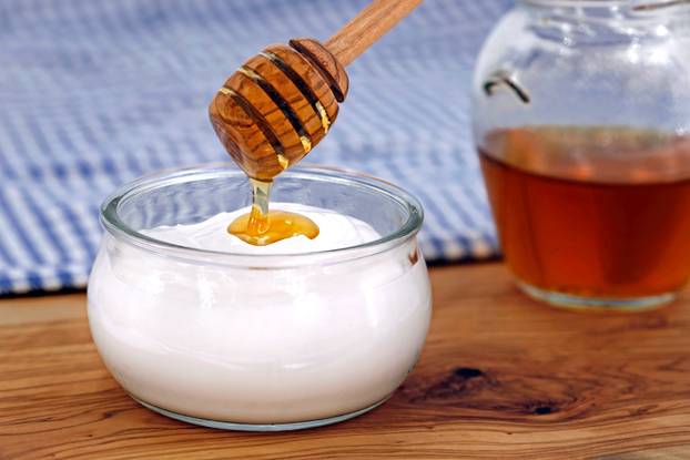 close up of wooden honey spoon pours honey on greek yogurt in glass jar on rustic olive wood table with traditional greek tablecloth in blue and white