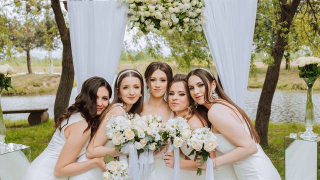 Group portrait of the bride and bridesmaids. Bride in a wedding dress and bridesmaids in white dresses and holding stylish bouquets on the wedding day against the background of the lake.