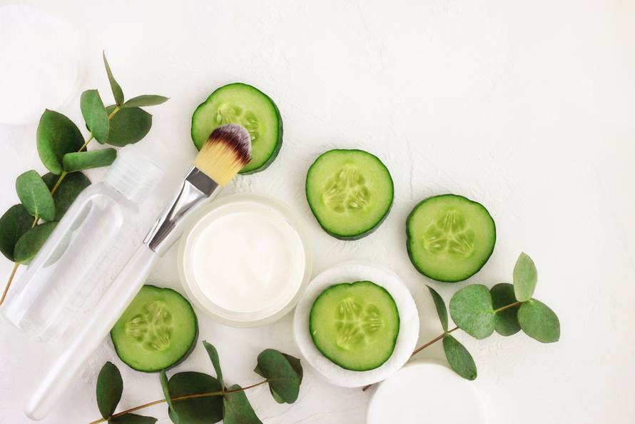 Cucumber slices, cosmetic cream jar and tonic mineral water in bottle, fresh green eucalyptus leaves, viewed above white background.