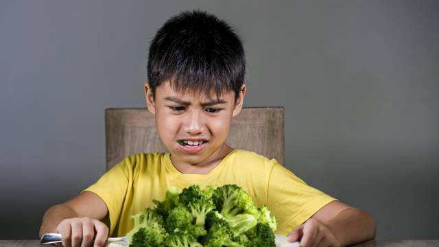 7 or 8 years old upset and disgusted Asian kid sitting on table in front of broccoli plate looking unhappy rejecting the fresh food in child hate green vegetables concept