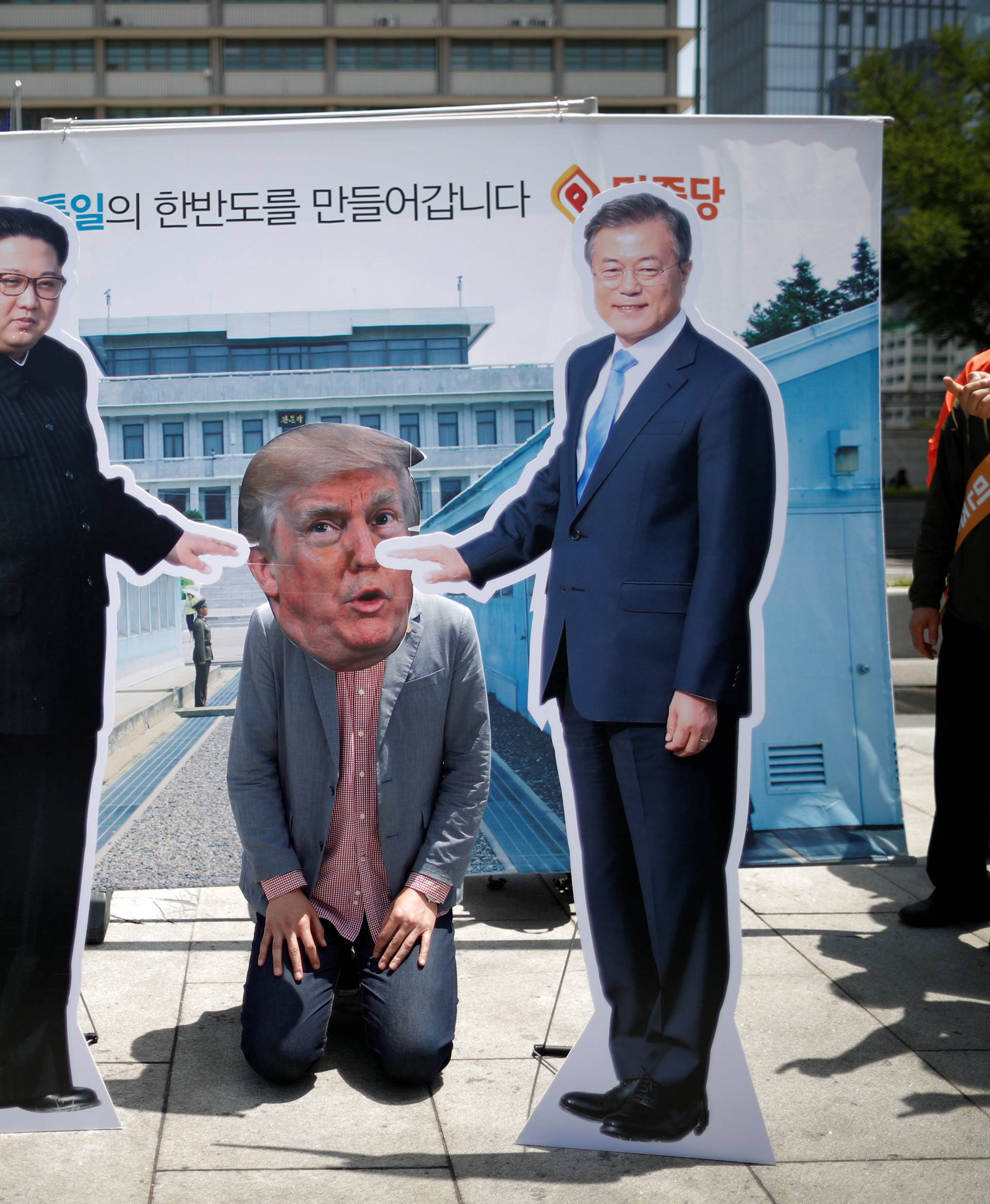 A man wearing a mask of U.S. President Donald Trump kneels down between cutouts of North Korean leader Kim Jong Un and South Korean President Moon Jae-in during an anti-U.S. President Donald Trump rally near U.S. embassy in Seoul