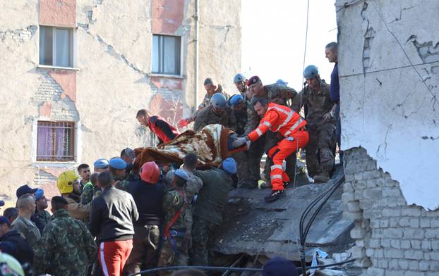 Military and emergency personnel carry an injured man on a stretcher near a damaged building in