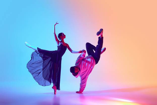 Ballet and street dance mix. Beautiful young girl in black dress, ballerina dancing with young man, hip hop performer on gradient background in neon