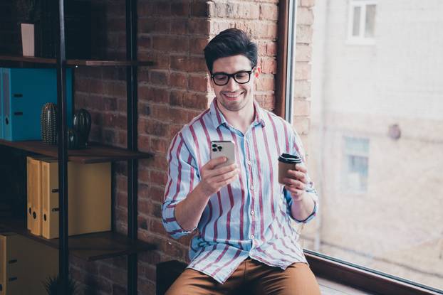Photo of nice young corporate man hold coffee smart phone wear striped shirt loft interior office indoors