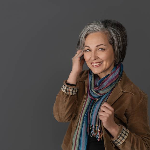 Creative mature woman in casual touching her graying short hair. Studio portrait on gray background. Copy space at left