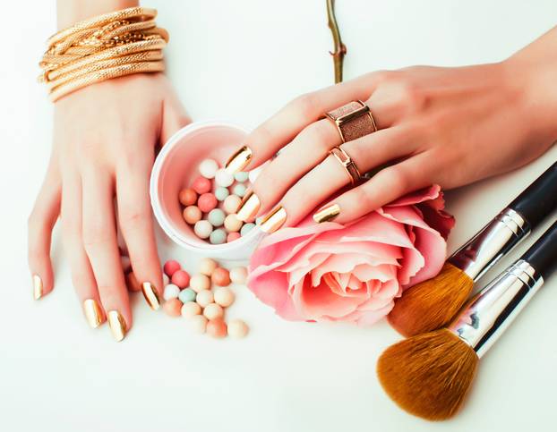 woman hands with golden manicure and many rings holding brushes, makeup artist stuff stylish, pure close up pink flower rose among cosmetic for makeup
