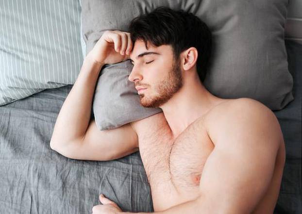 Single young muscular man sleeping in bed. He hold head on pillow. Young man is naked. Lower part of body covered with grey blanket.