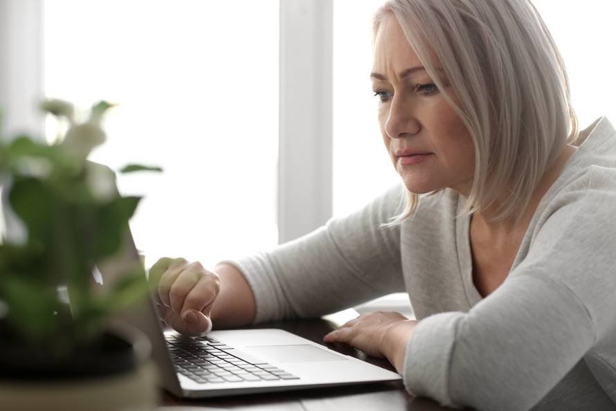 Mature,Woman,Reading,News,On,Laptop,Screen,In,Cafe