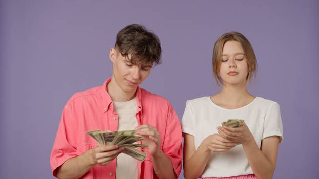 Positive couple of young boyfriend and girlfriend in pink white clothes isolated on purple background waving fan of money bills, spilling cash having fun smiling.